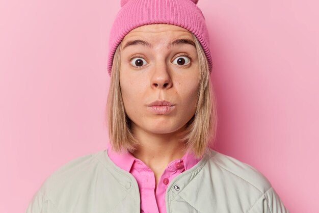 Horizontale Aufnahme einer schockierten Frau starrt verwanzte Augen an und bemerkt, dass etwas Beängstigendes nicht an schreckliche Nachrichten glauben kann, die in einem Jackenhut gekleidet sind, der isoliert über rosa Hintergrund gekleidet ist Konzept der menschlichen Reaktionen