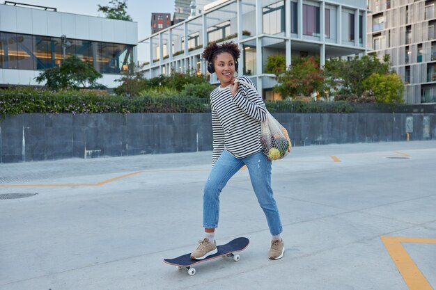 Horizontale Aufnahme eines glücklichen, lockigen, tausendjährigen Mädchens, das Skateboard in der Stadt reitet, gekleidet in Freizeitkleidung trägt Netztasche, hört Audiospur über drahtlose Kopfhörer schaut weg und lächelt freudig