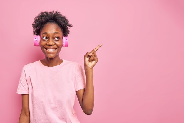 Horizontale Aufnahme eines optimistischen Teenager-Mädchens mit dunklem, positivem Gesichtsausdruck zeigt auf Kopienraum