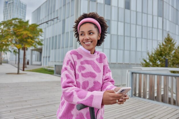 Horizontale Aufnahme eines Teenager-Mädchens mit lockigen Haaren, gekleidet in einem Pullover, fährt mit einem Elektroroller durch die Stadt, hält ein Mobiltelefon in den Händen, surft im Internet, liest Reisepublikationen in sozialen Medien und schaut weg