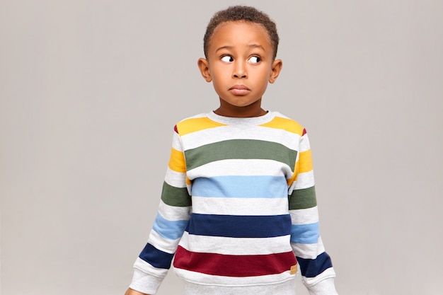 Horizontale Aufnahme eines verwirrten lustigen afroamerikanischen Jungen in einem gestreiften Pullover, der mit ahnungslosem Gesichtsausdruck wegschaut und sich schuldig fühlt, weil er alle Süßigkeiten aß und vorgab, unschuldig zu sein. Ich weiß es nicht
