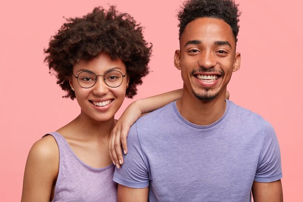 Horizontale Aufnahme von glücklichen afroamerikanischen Frau und Mann haben wahrheitsgemäße Beziehungen, zahniges Lächeln, glücklich, sich mit Freunden zu treffen, lässig gekleidet, isoliert über rosa Wand. Emotionskonzept