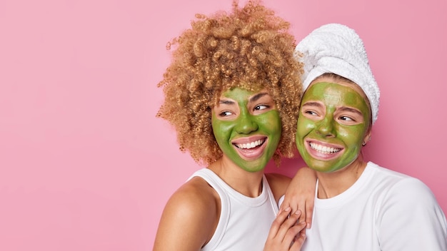 Horizontale Aufnahme von glücklichen Frauen wenden grüne Pflegemasken auf dem Gesicht an und schauen gerne weg, stehen dicht beieinander, isoliert über rosafarbenem Hintergrund, leerer Kopierraum für Ihre Werbeinhalte