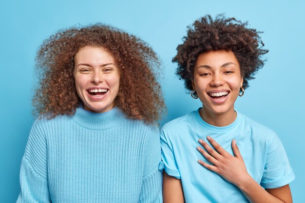 Horizontale Aufnahme von glücklichen, verschiedenen Frauen, die positiv kichern, fröhliche Ausdrücke stehen eng beieinander, drücken positive Emotionen aus, haben freundschaftliche Beziehungen isoliert über blauer Wand