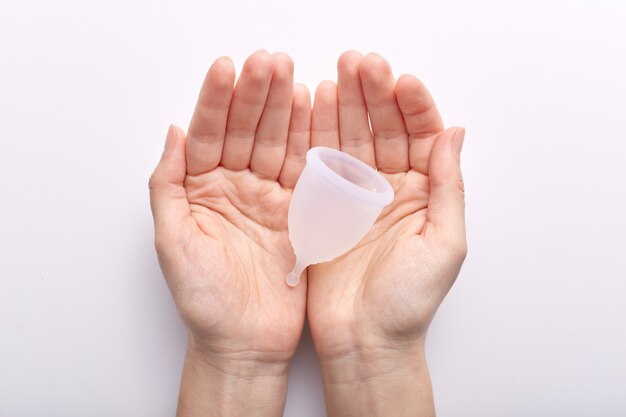 Horizontale Aufnahme von Händen, die sich über Weiß in Innenräumen zusammensetzen und saubere weiße Menstruationstasse halten