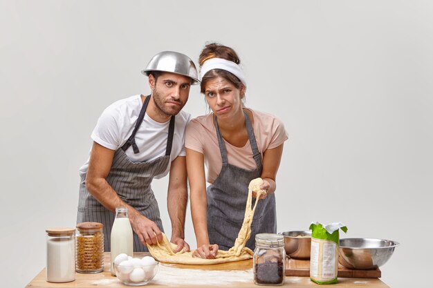 Horizontale Aufnahme von müder Hausfrau und Ehemann bereiten Teig mit Mehl zum Backen von Brot vor, probieren neues Rezept, müde vom Kochprozess, verbringen viele Stunden in der Küche. Vorbereitung für die Herstellung von frischem Gebäck