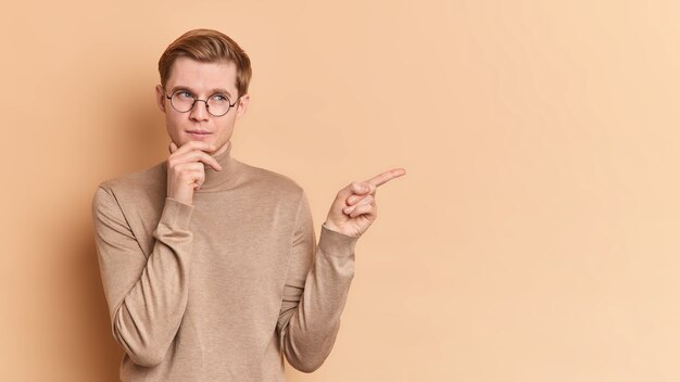 Horizontale Aufnahme von nachdenklichen jungen Mann hält Kinn denkt über vorgeschlagene Angebotspunkte entfernt zeigt Kopien Platz für Ihre Werbeinhalte trägt runde Brille Freizeitpullover