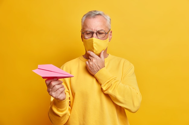 Horizontale Aufnahme von nachdenklichen männlichen Rentner in Brillen schaut aufmerksam auf Papierflugzeug hat ernsthafte Expession denkt, wie Krankheit zu überwinden trägt Schutzmaske während Quarantäne Posen in Innenräumen