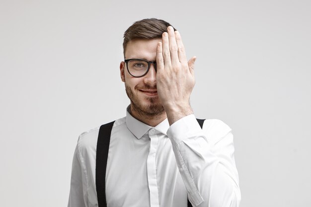 Horizontale Studioaufnahme eines glücklichen attraktiven jungen bärtigen Geschäftsmannes, der formelle Kleidung und Brille trägt, die eine Hälfte seines Gesichts bedeckt, während seine Augen bei Augenarzttermin untersucht werden