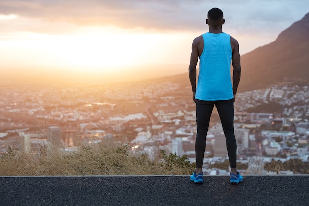 Horizontaler Panoramablick des nachdenklichen Sportlers in Sportkleidung tritt zurück, bewundert majestätische Berglandschaft und Sonnenaufgang, steht auf Asphalt hoch oben, Großstadt im Vordergrund.