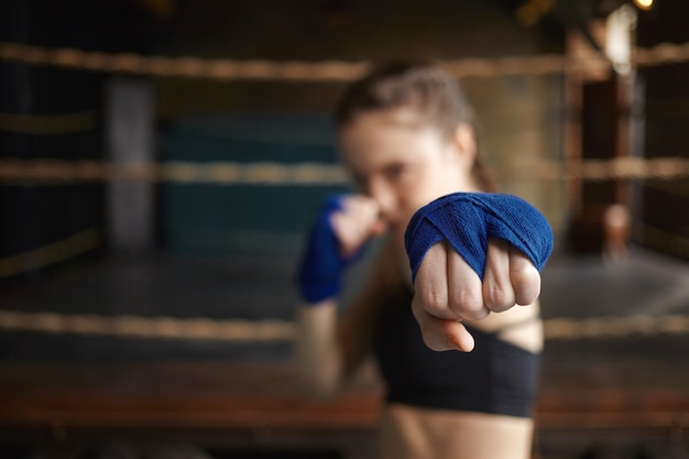 Horizontaler Schuss des stilvollen jungen Frauenboxers, der blaue Handwickel trägt, die drinnen trainieren, sich für den Boxkampf bereit machen und den Arm ausstrecken