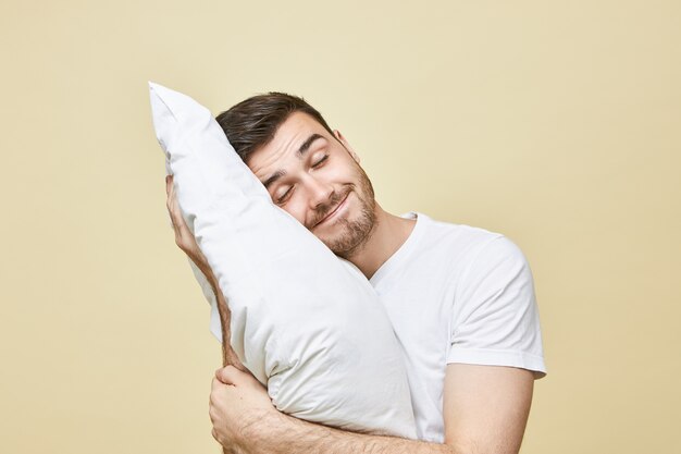 Horizontales Bild des hübschen niedlichen jungen Mannes mit der Borste, die mit Kopf auf weißem weichem Kissen aufwirft, das friedlich schläft und lächelt, guten Traum seeling. Attraktives Nickerchen nach einem harten Arbeitstag