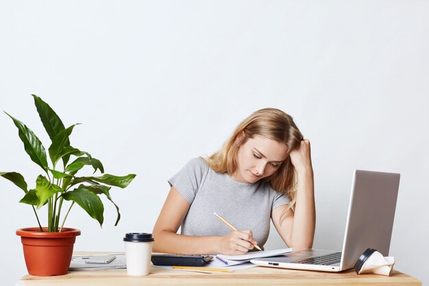 Horizontales Porträt der ernsthaften blonden jungen Geschäftsfrau, die mit Bleistift in ihrem Notizbuch schaut, Zahlen berechnet, Jahresbericht für ihre eigene Firma macht, Laptop für ihre Arbeit verwendend