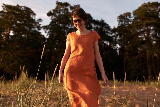 Horizontales Porträt der fröhlichen jungen brünetten Frau, die Sonnenbrille und rotes Baumwollkleid trägt, das draußen mit freudigem Lächeln aufwirft