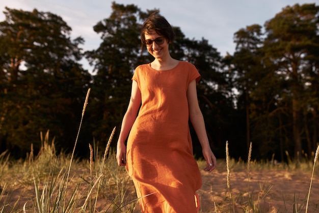 Horizontales Porträt der fröhlichen jungen brünetten Frau, die Sonnenbrille und rotes Baumwollkleid trägt, das draußen mit freudigem Lächeln aufwirft