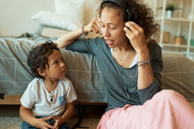 Horizontales Porträt der schönen jungen hispanischen Frau, die Musik in kabellosen Kopfhörern hört, die auf Boden mit ihrem hübschen kleinen Sohn sitzen. Glückliche Familie, die Spaß zu Hause hat