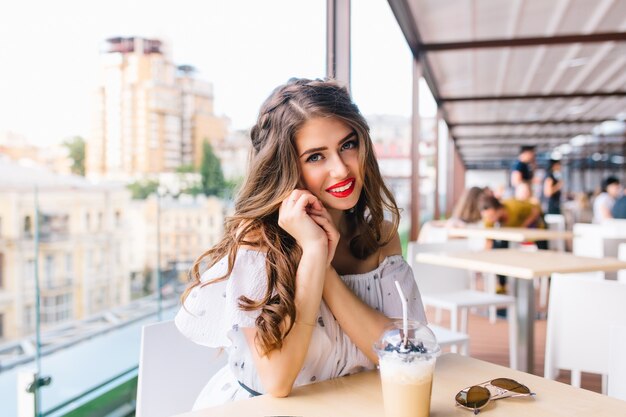 Horizontales Porträt des hübschen Mädchens mit dem langen Haar, das am Tisch auf der Terrasse im Café sitzt. Sie trägt ein weißes Kleid mit nackten Schultern und rotem Lippenstift. Sie lächelt freundlich in die Kamera.