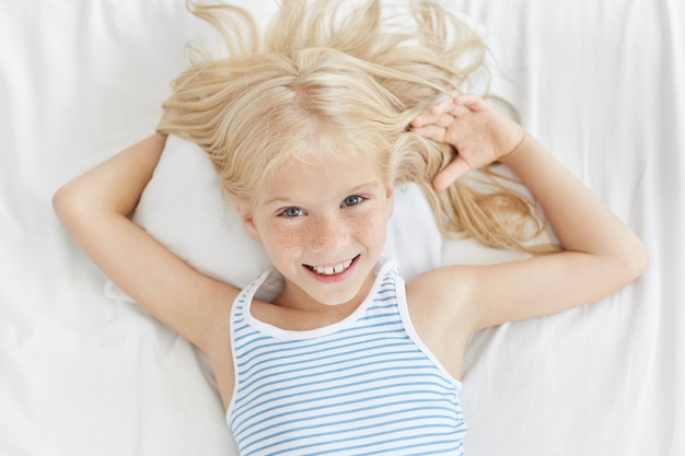 Horizontales Porträt des niedlichen Mädchens mit blau leuchtenden Augen, sommersprossigem Gesicht und sanftem Lächeln, entspannend im Bett, liegend auf bequemem weißem Kissen