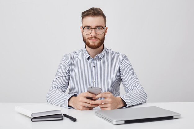 Horizontales Porträt des unrasierten angenehm aussehenden jungen Geschäftsmannes sitzt am Schreibtisch,