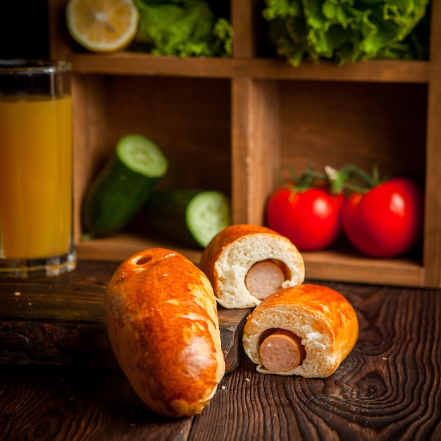 Hot Dog mit Orangensaft und Gurken sowie Tomaten und Salat in Holzbrett