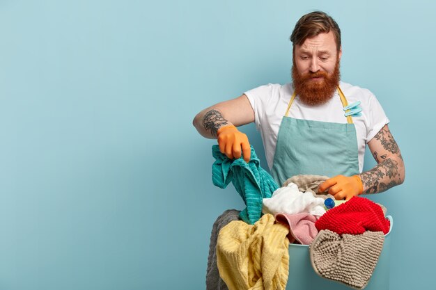 Housekeeping- und Hausarbeitskonzept. Frustrierter bärtiger bärtiger Mann hält Handtuch, wählt schmutzige Wäsche aus dem Korb