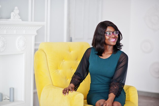 Hübsche afrikanisch-amerikanische Frau mit Brille posiert im Zimmer auf gelbem Stuhl sitzend