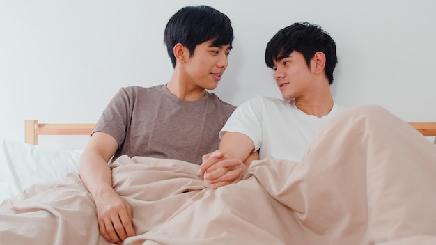 Hübsche asiatische homosexuelle Paare, die zu Hause auf Bett sprechen. Der junge asiatische glückliche LGBTQ + Kerl entspannen sich, verbringen zusammen romantische Zeit, nachdem er morgens im Schlafzimmer am modernen Haus aufgewacht ist.