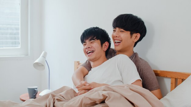 Hübsche asiatische homosexuelle Paare, die zu Hause auf Bett sprechen. Der junge asiatische glückliche LGBTQ + Kerl entspannen sich, verbringen zusammen romantische Zeit, nachdem er morgens im Schlafzimmer am modernen Haus aufgewacht ist.