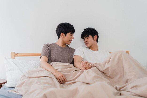 Hübsche asiatische homosexuelle Paare, die zu Hause auf Bett sprechen. Der junge asiatische glückliche LGBTQ + Kerl entspannen sich, verbringen zusammen romantische Zeit, nachdem er morgens im Schlafzimmer am modernen Haus aufgewacht ist.