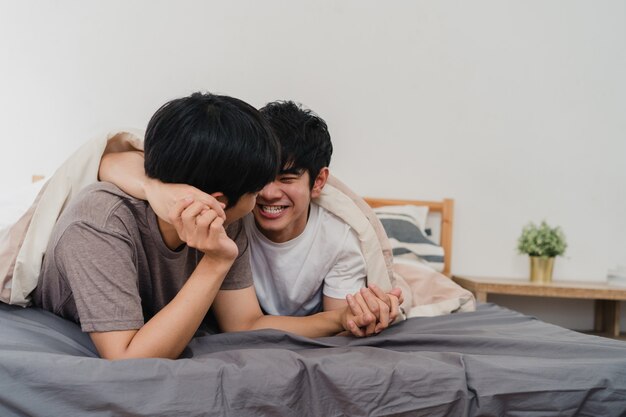 Hübsche asiatische homosexuelle Paare, die zu Hause auf Bett sprechen. Der junge asiatische glückliche LGBTQ + Kerl entspannen sich, verbringen zusammen romantische Zeit, nachdem er morgens im Schlafzimmer am modernen Haus aufgewacht ist.
