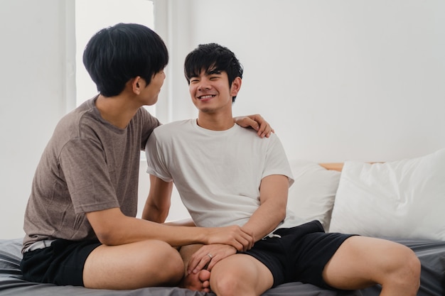 Hübsche asiatische homosexuelle Paare, die zu Hause auf Bett sprechen. Der junge asiatische glückliche LGBTQ + Kerl entspannen sich, verbringen zusammen romantische Zeit, nachdem er morgens im Schlafzimmer am modernen Haus aufgewacht ist.