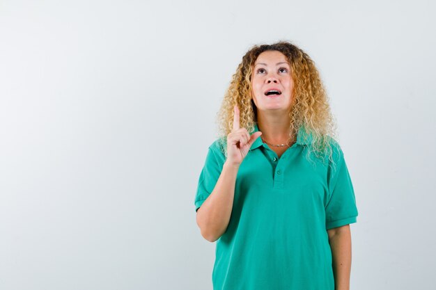 Hübsche blonde Dame im grünen Polo-T-Shirt zeigt nach oben, schaut nach oben und schaut dankbar, Vorderansicht.