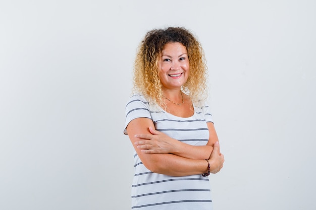 Hübsche blonde Frau, die die Arme im gestreiften T-Shirt verschränkt hält und froh aussieht, Vorderansicht.