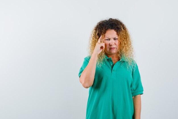 Hübsche blonde Frau, die Finger auf Schläfen im grünen Polo-T-Shirt hält und nachdenklich aussieht. Vorderansicht.