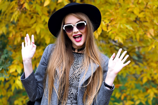 Hübsche blonde Frau, die Spaß am Stadtpark im kühlen Herbsttag, elegantes trendiges Outfit, Schal, Hutsonnenbrille, Halsband, eleganter Straßenstil hat