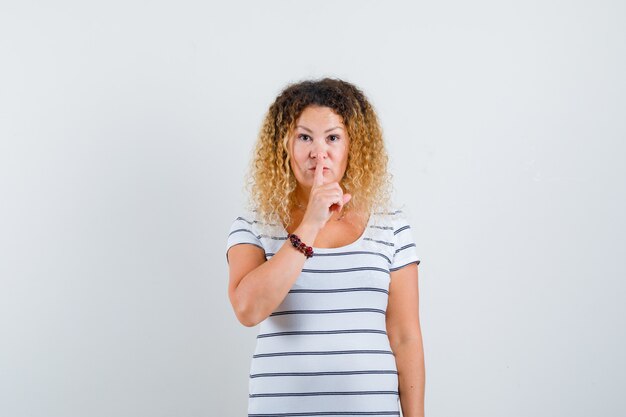 Hübsche blonde Frau, die Stillegeste in gestreiftem T-Shirt zeigt und vorsichtig aussieht, Vorderansicht.