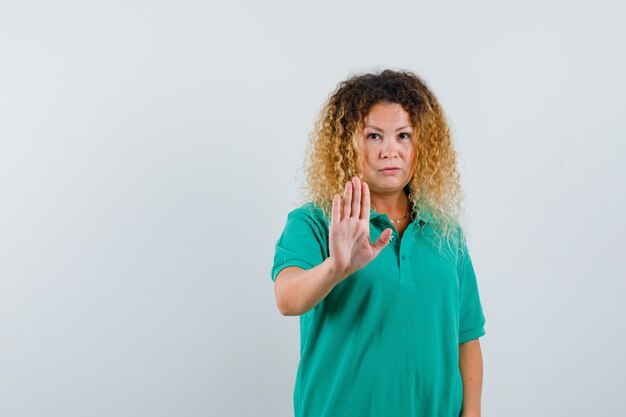 Hübsche blonde Frau, die Stoppschild im grünen Polo-T-Shirt zeigt und ernst schaut. Vorderansicht.