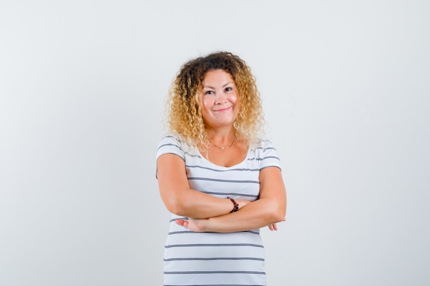 Hübsche blonde Frau im gestreiften T-Shirt, die die Arme verschränkt hält und fröhlich aussieht, Vorderansicht.