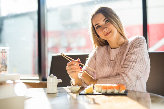 Hübsche blonde Frau im weißen Pullover, die Sushi zum Mittagessen an einem kleinen Kaffee isst