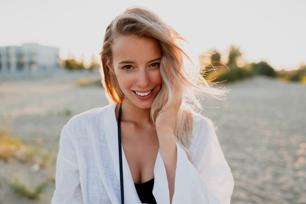 Hübsche blonde Frau in der weißen Bluse, die am Strand aufwirft. Sommerstimmung. Tropischer Urlaub. Windige Haare.
