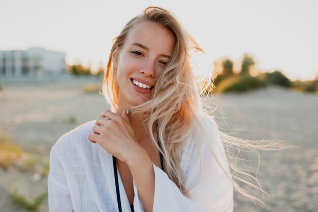 Hübsche blonde Frau in der weißen Bluse, die am Strand aufwirft. Sommerstimmung. Tropischer Urlaub. Windige Haare.