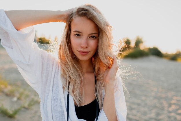 Hübsche blonde Frau in der weißen Bluse, die am Strand aufwirft. Sommerstimmung. Tropischer Urlaub.