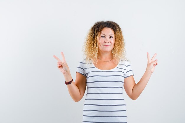 Hübsche blonde Frau in gestreiftem T-Shirt, die Friedensgeste zeigt und positiv aussieht, Vorderansicht.