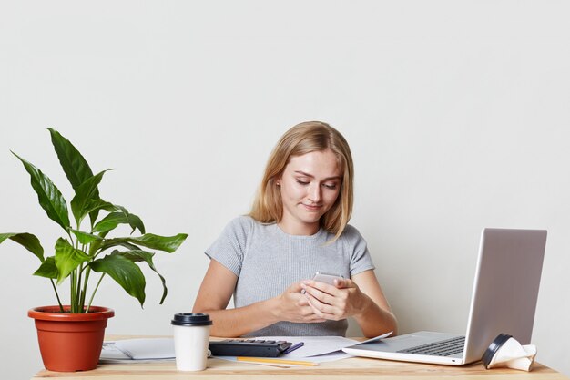 Hübsche blonde junge Unternehmerin sitzt am Arbeitsplatz, erstellt einen Jahresbericht für ein Geschäftstreffen, hält ein zeitgemäßes Mobiltelefon in der Hand und ruft den Partner an, um Beratung oder guten Rat zu erhalten