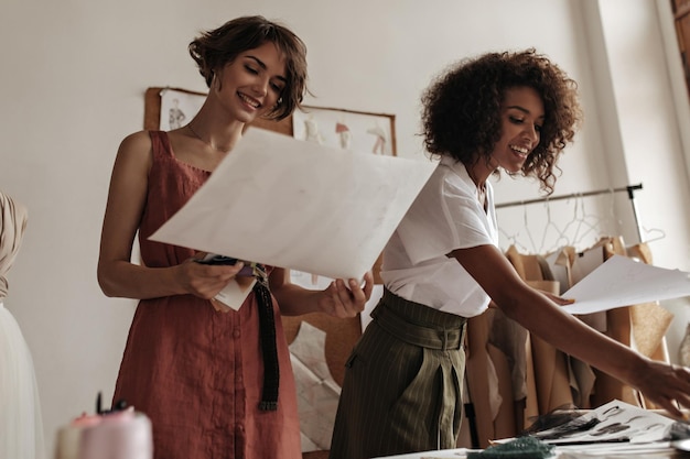 Hübsche, brünette, attraktive junge Damen in trendigen Outfits lächeln, arbeiten als Modedesigner und halten große Papierbögen mit bemalten Kleidern