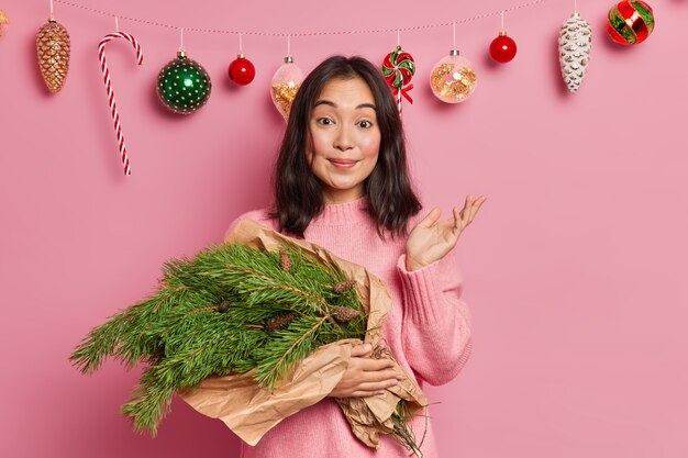 Hübsche brünette junge Frau hat kreatives Hobby hebt Palme mit Zögern nicht weiß nicht, was man aus grünen Fichtenzweigen macht, bereitet sich auf Winterferien vor Posen über Weihnachtsdekor zu Hause