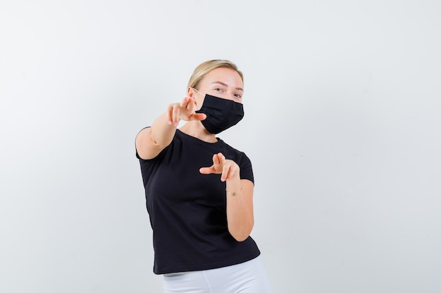 Hübsche Dame, die in schwarzem T-Shirt auf die Kamera zeigt, schwarze Maske isoliert