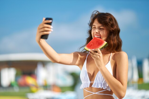 Hübsche Dame im Bikini macht Selfie, während sie im Urlaub reife Wassermelone beißt