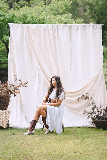 Hübsche Dame im langen weißen Kleid mit Gürtel, der im Garten mit weißer Wand während des Tages sitzt und lächelt.