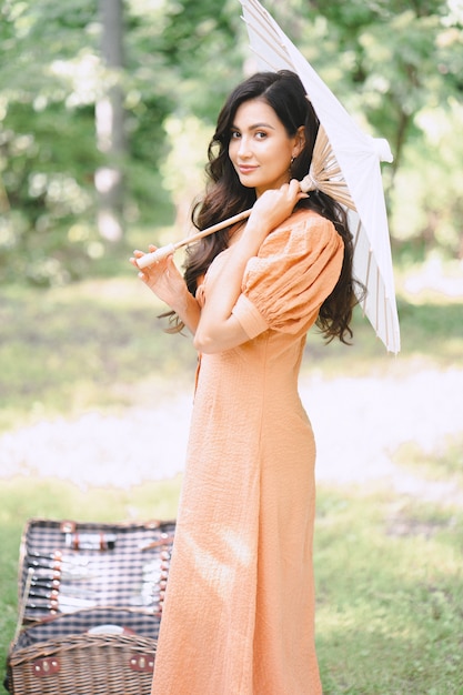 Hübsche Dame im orangefarbenen Kleid, das Regenschirm hält und tagsüber in der Natur schaut.